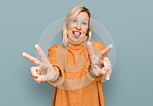 Middle age caucasian woman wearing casual winter sweater smiling with tongue out showing fingers of both hands doing victory sign