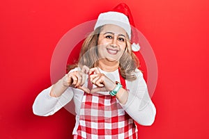 Middle age caucasian woman wearing cook apron and christmas hat smiling in love showing heart symbol and shape with hands