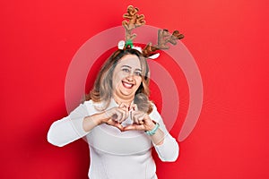 Middle age caucasian woman wearing cute christmas reindeer horns smiling in love doing heart symbol shape with hands
