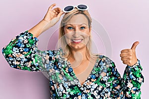 Middle age caucasian woman wearing floral shirt and sunglasses smiling happy and positive, thumb up doing excellent and approval