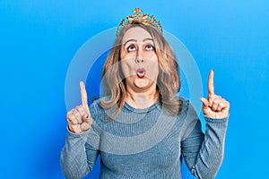 Middle age caucasian woman wearing queen crown amazed and surprised looking up and pointing with fingers and raised arms