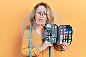 Middle age caucasian woman wearing tape measure holding sew kit in shock face, looking skeptical and sarcastic, surprised with