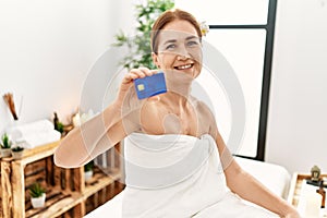 Middle age caucasian woman wearing towel holding credit card sitting on massage board at beauty center