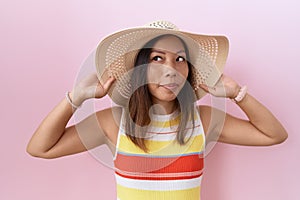 Middle age chinese woman wearing summer hat over pink background smiling looking to the side and staring away thinking