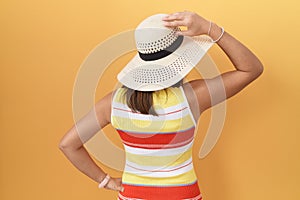 Middle age chinese woman wearing summer hat over yellow background backwards thinking about doubt with hand on head