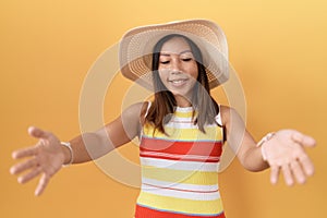 Middle age chinese woman wearing summer hat over yellow background looking at the camera smiling with open arms for hug