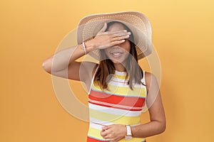 Middle age chinese woman wearing summer hat over yellow background smiling and laughing with hand on face covering eyes for