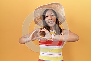 Middle age chinese woman wearing summer hat over yellow background smiling in love doing heart symbol shape with hands