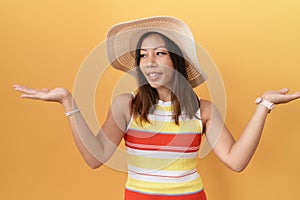 Middle age chinese woman wearing summer hat over yellow background smiling showing both hands open palms, presenting and