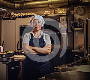 A middle age cook wearing a uniform standing with her arms crossed at restaurant`s kitchen.