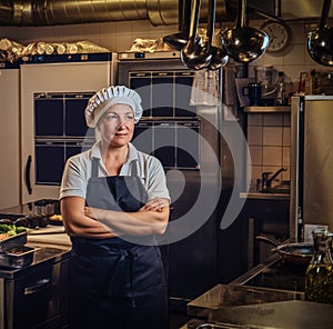 A middle age cook wearing a uniform standing with her arms crossed at restaurant`s kitchen.