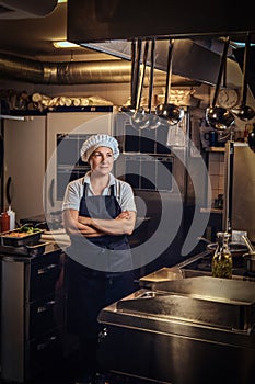 A middle age cook wearing a uniform standing with her arms crossed at restaurant`s kitchen.