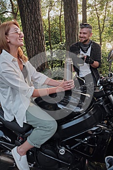 Middle age couple drinking tea, sitting on a motorcycle, traveling together on a forest road