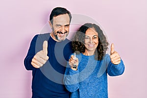 Middle age couple of hispanic woman and man holding keys of new home approving doing positive gesture with hand, thumbs up smiling