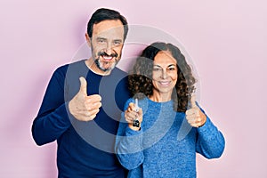 Middle age couple of hispanic woman and man holding keys of new home doing happy thumbs up gesture with hand