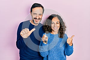 Middle age couple of hispanic woman and man holding keys of new home pointing to the back behind with hand and thumbs up, smiling