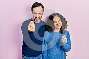 Middle age couple of hispanic woman and man hugging and standing together doing money gesture with hands, asking for salary