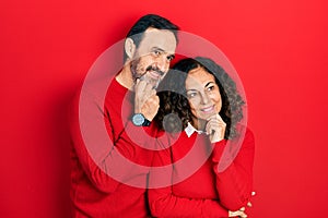 Middle age couple of hispanic woman and man hugging and standing together with hand on chin thinking about question, pensive