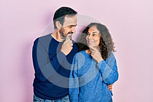 Middle age couple of hispanic woman and man hugging and standing together with hand on chin thinking about question, pensive