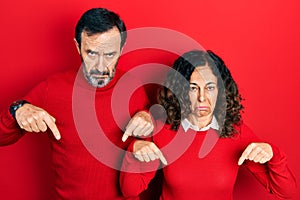 Middle age couple of hispanic woman and man hugging and standing together pointing down looking sad and upset, indicating