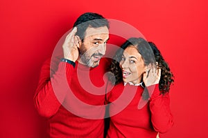 Middle age couple of hispanic woman and man hugging and standing together smiling with hand over ear listening an hearing to rumor