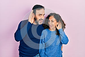 Middle age couple of hispanic woman and man hugging and standing together smiling with hand over ear listening an hearing to rumor