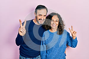 Middle age couple of hispanic woman and man hugging and standing together smiling looking to the camera showing fingers doing