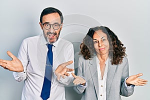 Middle age couple of hispanic woman and man wearing business office uniform clueless and confused expression with arms and hands