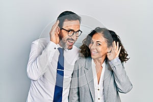 Middle age couple of hispanic woman and man wearing business office uniform smiling with hand over ear listening an hearing to