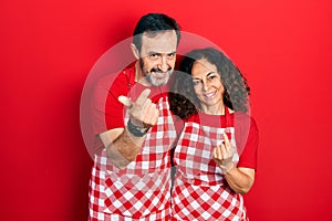 Middle age couple of hispanic woman and man wearing cook apron beckoning come here gesture with hand inviting welcoming happy and