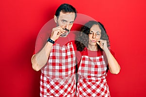 Middle age couple of hispanic woman and man wearing cook apron mouth and lips shut as zip with fingers