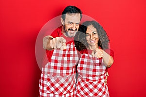 Middle age couple of hispanic woman and man wearing cook apron pointing to you and the camera with fingers, smiling positive and