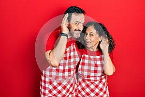 Middle age couple of hispanic woman and man wearing cook apron smiling with hand over ear listening an hearing to rumor or gossip