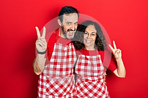 Middle age couple of hispanic woman and man wearing cook apron smiling looking to the camera showing fingers doing victory sign