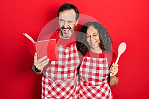 Middle age couple of hispanic woman and man wearing professional apron reading cooking recipe book winking looking at the camera