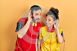 Middle age couple of hispanic woman and man wearing sportswear and arm band smiling with hand over ear listening an hearing to