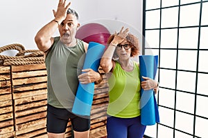 Middle age couple holding yoga mat surprised with hand on head for mistake, remember error