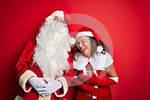 Middle age couple wearing Santa costume and glasses over isolated red background smiling and laughing hard out loud because funny