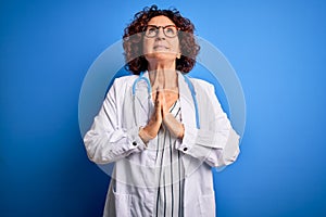 Middle age curly hair doctor woman wearing coat and stethoscope over blue background begging and praying with hands together with