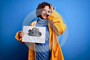 Middle age curly hair woman wearing rain coat holding banner with cloud over blue background with happy face smiling doing ok sign