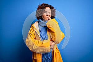 Middle age curly hair woman wearing rain coat standing over isolated blue background with hand on chin thinking about question,