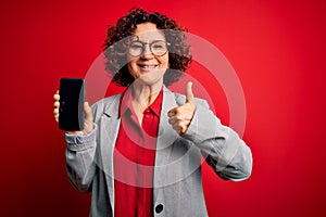 Middle age curly woman holding smartphone showing screen over  red background happy with big smile doing ok sign, thumb up