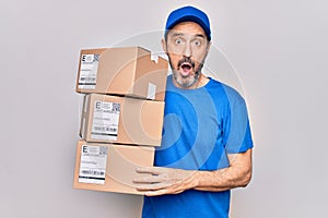 Middle age delivery man wearing cap holding package over isolated white background scared and amazed with open mouth for surprise,
