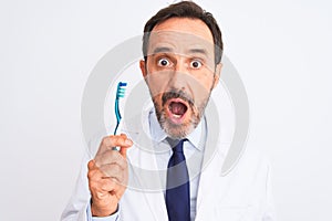 Middle age dentist man holding toothbrush standing over isolated white background scared in shock with a surprise face, afraid and