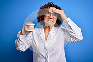 Middle age dentist woman wearing coat holding plastic denture teeth over blue background stressed with hand on head, shocked with