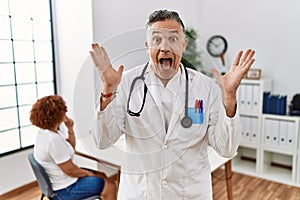 Middle age doctor man at the clinic with a patient celebrating crazy and amazed for success with arms raised and open eyes