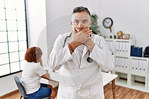 Middle age doctor man at the clinic with a patient shocked covering mouth with hands for mistake