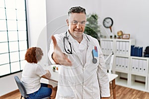 Middle age doctor man at the clinic with a patient smiling friendly offering handshake as greeting and welcoming