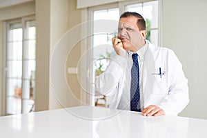 Middle age doctor man wearing medical coat at the clinic looking stressed and nervous with hands on mouth biting nails