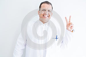 Middle age doctor man wearing medical coat over white background smiling with happy face winking at the camera doing victory sign